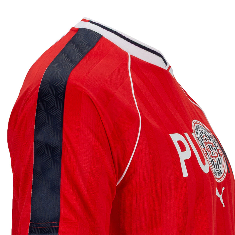 Red soccer jersey shoulder close-up with navy panel, white piping, and crest detail.