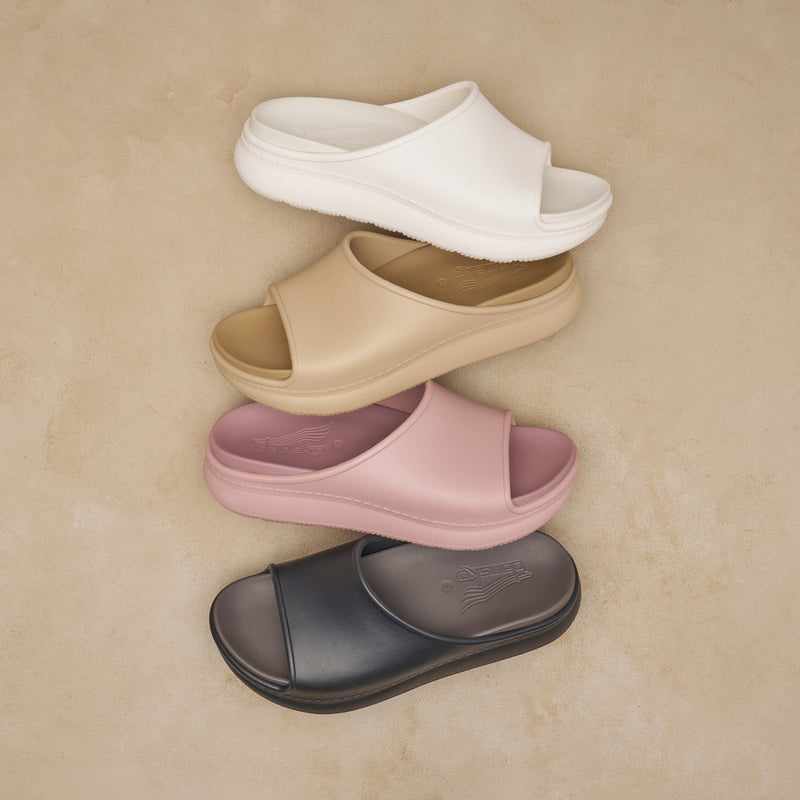 Flat lay of a recovery slide in four colorways: white, tan, pink and black. They are shot on a neutral backdrop.